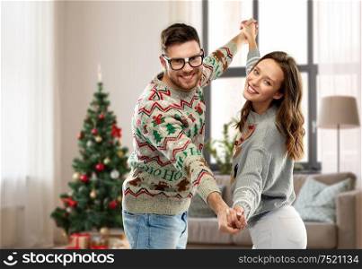 celebration, fun and holidays concept - happy couple wearing ugly sweaters dancing over christmas tree and home background. couple dancing in christmas ugly sweaters at home