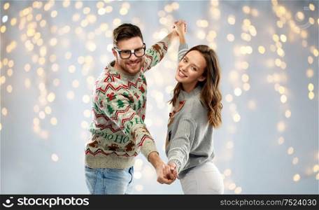 celebration, fun and holidays concept - happy couple wearing ugly sweaters dancing at christmas party over festive lights background. couple dancing at christmas ugly sweaters party