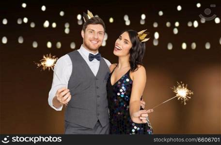 celebration, fun and holidays concept - happy couple with crowns and sparklers at party over garland lights background. happy couple with crowns and sparklers at party