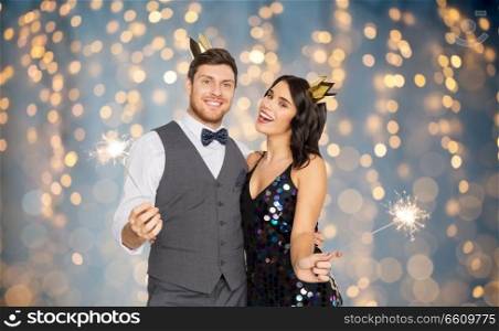 celebration, fun and holidays concept - happy couple with crowns and sparklers at party over festive lights background. happy couple with crowns and sparklers at party