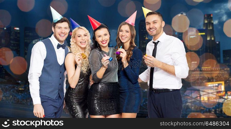 celebration, fun and holidays concept - happy friends with party blowers and caps over night singapore city and lights background. happy friends with party blowers having fun. happy friends with party blowers having fun