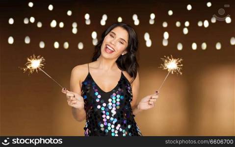 celebration, fun and holidays concept - happy young woman in sequin dress with sparklers at party over garland lights background. happy young woman with sparklers at party