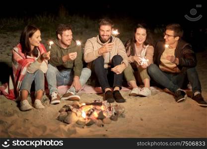 celebration, holidays and people concept - happy friends with sparklers sitting at camp fire on beach at night. happy friends with sparklers at camp fire at night