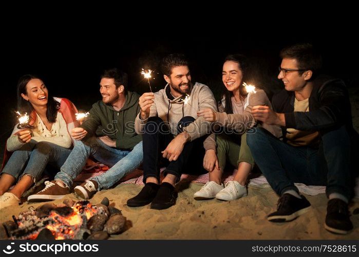 celebration, holidays and people concept - happy friends with sparklers sitting at camp fire at night. happy friends with sparklers at camp fire at night