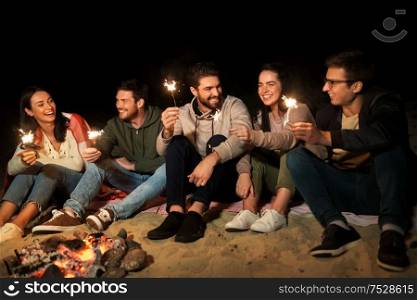 celebration, holidays and people concept - happy friends with sparklers sitting at camp fire at night. happy friends with sparklers at camp fire at night