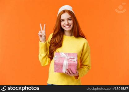 Celebration, holidays and presents concept. Cute cheerful redhead female in winter hat, sweater, holding pink present, showing peace sign as receive gift and smiling delighted, orange background.. Celebration, holidays and presents concept. Cute cheerful redhead female in winter hat, sweater, holding pink present, showing peace sign as receive gift and smiling delighted, orange background