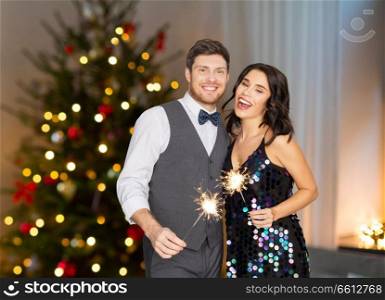celebration, party and holidays concept - happy couple with sparklers over christmas tree lights background. happy couple with sparklers at christmas party