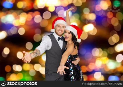 celebration, people and holidays concept - happy couple in santa hats at christmas or new year party over festive lights background. happy couple in santa hats at christmas party