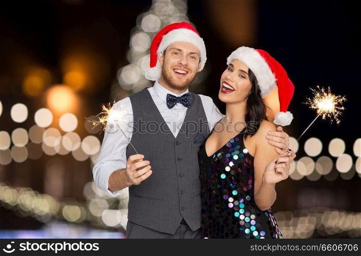 celebration, people and holidays concept - happy couple in santa hats at christmas or new year party with sparklers over lights background. happy couple with sparklers at christmas party