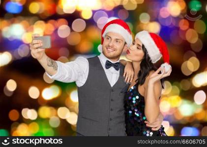 celebration, people and holidays concept - happy couple in santa hats taking selfie by smartphone at christmas or new year party over festive lights background. couple in santa hats taking selfie at christmas