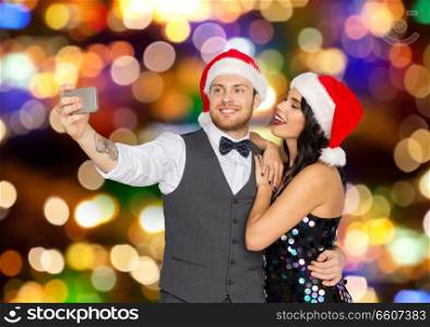 celebration, people and holidays concept - happy couple in santa hats taking selfie by smartphone at christmas or new year party over festive lights background. couple in santa hats taking selfie at christmas