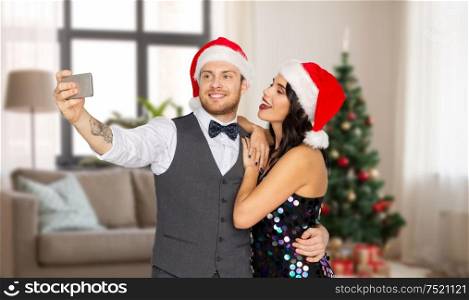 celebration, people and holidays concept - happy couple in santa hats taking selfie by smartphone over christmas tree on home background. couple in santa hats taking selfie on christmas