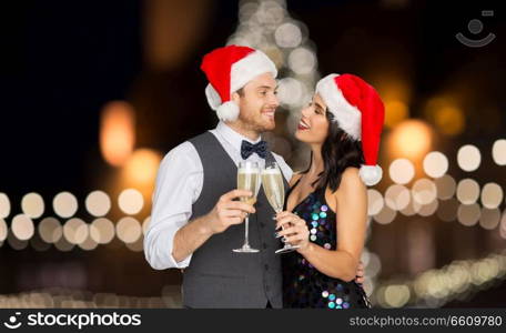 celebration, people and holidays concept - happy couple in santa hats with glasses of non alcoholic champagne over christmas tree lights background. couple with champagne glasses at christmas party