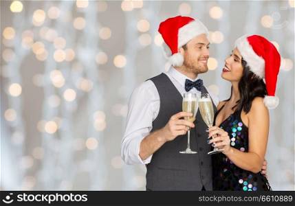 celebration, people and holidays concept - happy couple in santa hats with glasses of non alcoholic champagne at christmas or new year party over festive lights background. couple with champagne glasses at christmas party