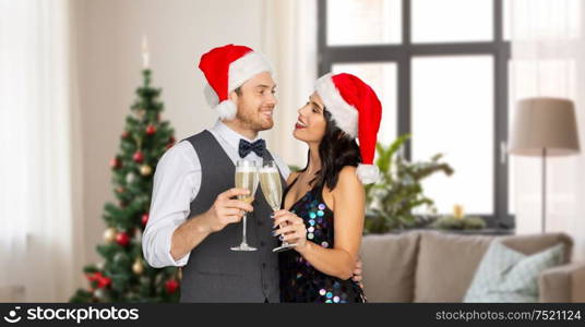 celebration, people and holidays concept - happy couple in santa hats with glasses of non alcoholic champagne over christmas tree on home background. couple with champagne glasses at home on christmas