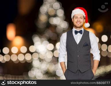 celebration, people and holidays concept - happy man in santa hat and suit over christmas tree lights background. happy man in santa hat and suit at christmas