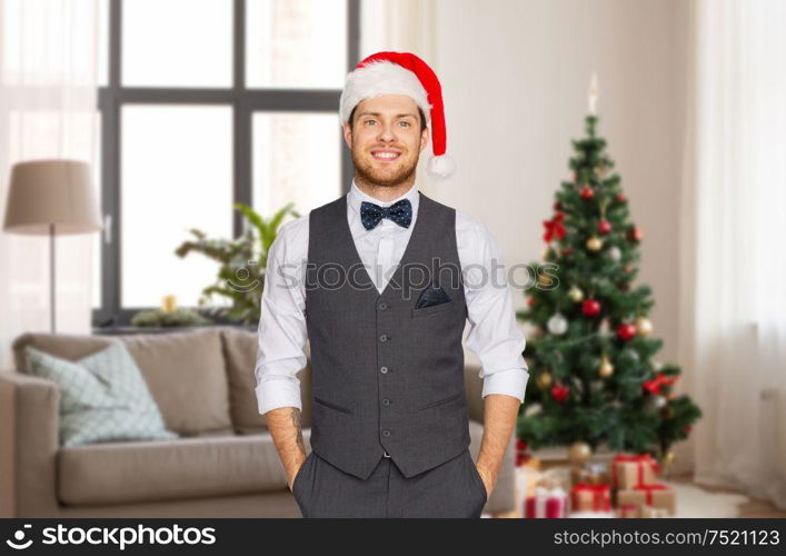 celebration, people and holidays concept - happy man in santa hat and suit over christmas tree on home background. man in santa hat and suit at home on christmas