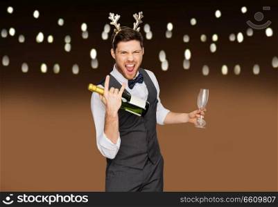 celebration, people and holidays concept - happy man with bottle of champagne and wine glasses at christmas or new year party making rock gesture over garland lights background. man with bottle of champagne at christmas party