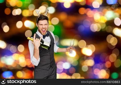 celebration, people and holidays concept - happy man with bottle of champagne and wine glasses at christmas or new year party making rock gesture over festive lights background. man with bottle of champagne at christmas party