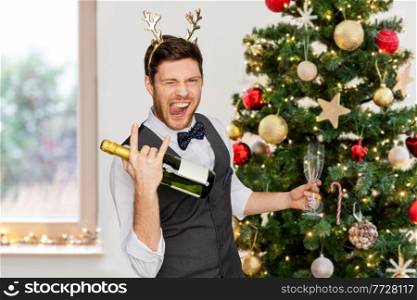 celebration, people and holidays concept - happy man with bottle of champagne and wine glasses at party making rock gesture over christmas tree on background. man with bottle of champagne at christmas party