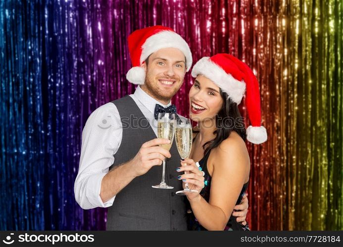 celebration, people and winter holidays concept - happy couple in santa hats with glasses of non alcoholic champagne at christmas party over foil fringe on background. couple with champagne glasses at christmas party