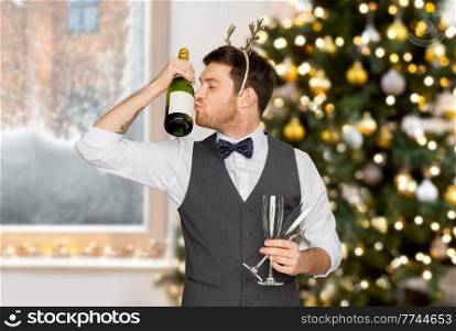 celebration, people and winter holidays concept - happy man kissing bottle of champagne and wine glasses at party over christmas tree lights on background. man kissing bottle of champagne at christmas party