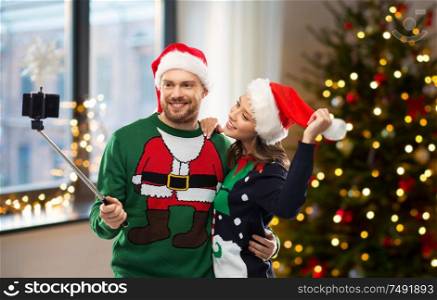 celebration, technology and winter holidays concept - happy couple in santa hats taking picture by smartphone on selfie stick at ugly sweater party over christmas tree at home background. happy couple in christmas sweaters taking selfie