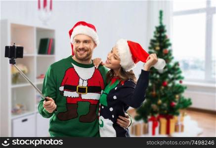 celebration, technology and winter holidays concept - happy couple in santa hats taking picture by smartphone on selfie stick at ugly sweater party over christmas tree at home background. happy couple in christmas sweaters taking selfie