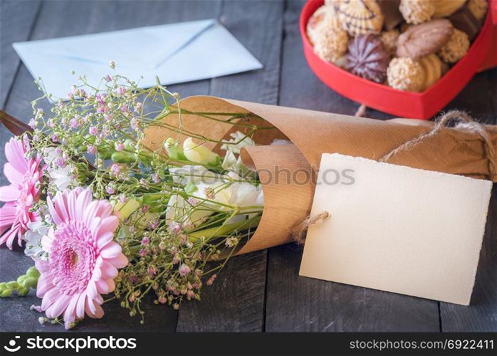 Celebration theme image with a lovely bouquet of flowers wrapped in brown paper with a tag attached to it, surrounded by a box of cookies and an envelope.