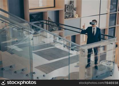 CEO director of gaming company testing prototype of VR headset glasses while walking around office center lobby and imitating movements in virtual reality, gesturing with index finger up in air. CEO of gaming company testing prototype of VR headset while walking around office center lobby