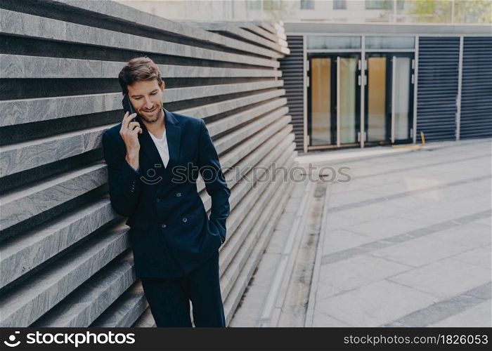 CEO male in smart casual having pleasing phone talk standing outside of office center entrance, leans on marbale striped facing outer wall, having coversation on mobile about upcoming board meeting. CEO male in having pleasing phone talk on mobile while standing outside of office center entrance
