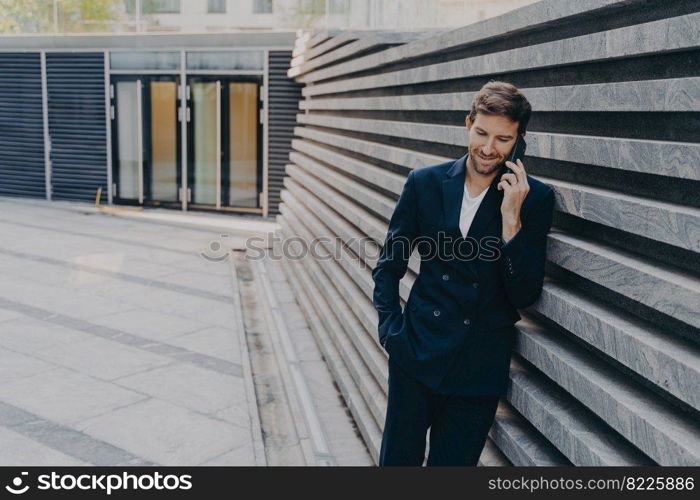 CEO male in smart casual having pleasing phone talk standing outside of office center entrance, leans on marbale striped facing outer wall, having coversation on mobile about upcoming board meeting. CEO male in having pleasing phone talk on mobile while standing outside of office center entrance