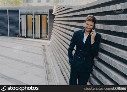 CEO male in smart casual having pleasing phone talk standing outside of office center entrance, leans on marbale striped facing outer wall, having coversation on mobile about upcoming board meeting. CEO male in having pleasing phone talk on mobile while standing outside of office center entrance