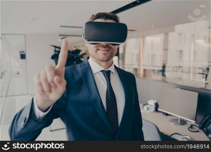 CEO man stands in office with VR headset and interacts with application virtual interface, pressing invisible buttons or selects menu items moving his forefinger in air. Cyberspace experience concept. CEO man stands in office with VR headset and interacts with application virtual interface