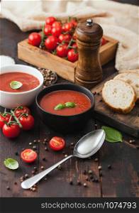 Ceramic bowl plates of creamy tomato soup with spoon, pepper and kitchen cloth on wooden board with box of raw tomatoes and bread.