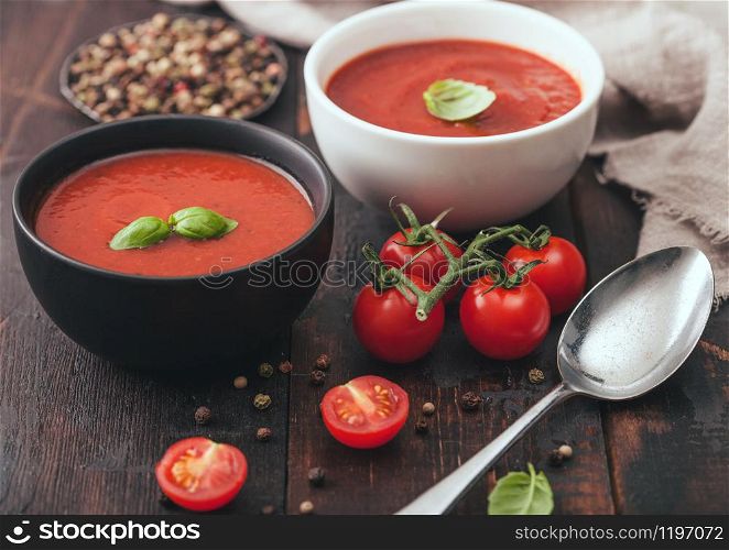 Ceramic bowl plates of creamy tomato soup with spoon, pepper and kitchen cloth on wooden board with raw tomatoes.
