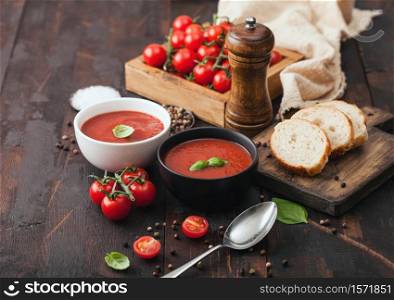 Ceramic bowl plates of creamy tomato soup with spoon, pepper and kitchen cloth on wooden board with box of raw tomatoes and bread.