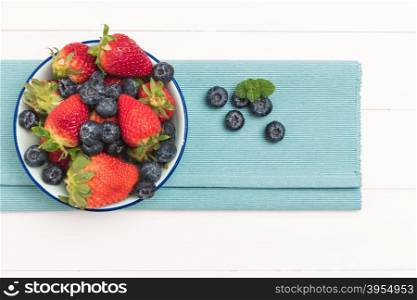 Ceramic bowl with blueberries and strawberries at blue textile napkin over wooden table. Top view with copy space