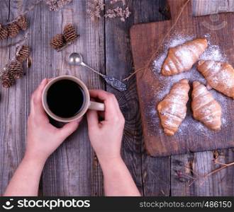 ceramic brown cup with black coffee and wooden cutting board with baked croissants, baked with icing sugar, hands holding a mug
