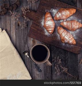 ceramic brown cup with black coffee and wooden cutting board with baked croissants, baked with powdered sugar, top view