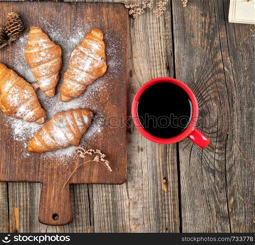 ceramic red cup with black coffee and wooden cutting board with baked croissants, baked with powdered sugar, top view