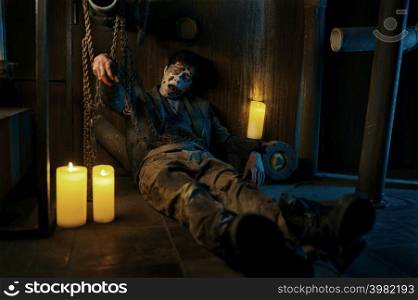 Chained zombie surrounded with candle sitting in basement of abandoned house. Mysterious nightlife. Chained zombie in basement of abandoned house