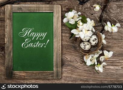 chalkboard, flowers and easter nest with eggs on rustic wooden background with sample text Happy Easter!