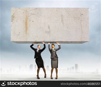Challenge in business. Image of two businesswomen holding stone above head. Partnership and cohesion