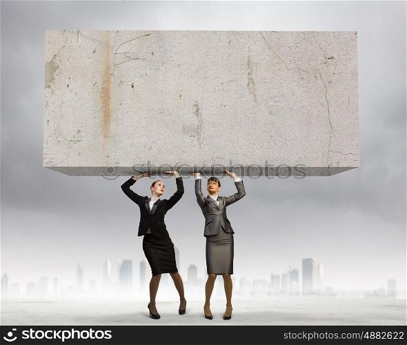 Challenge in business. Image of two businesswomen holding stone above head. Partnership and cohesion
