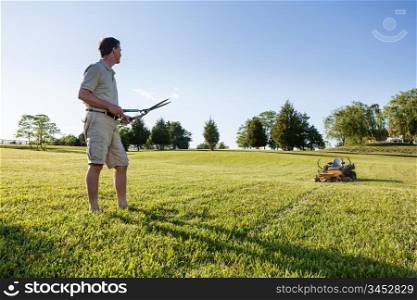 Challenging task of cutting large lawn with grass shears by hand with mower in background