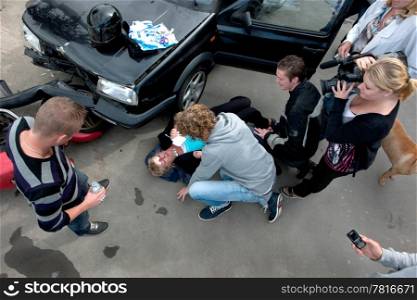 Chaotic scene just after a car crash, with an injured woman lying on the ground, several bystanders providing first aid, and a television reporter with camera capturing the scene, seen from above