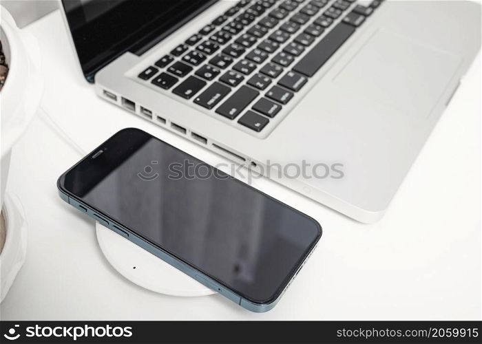 Charging smartphone battery with wireless charger device near laptop computer, Modern technology transfer energy mobile phone on workspace desktop at home office