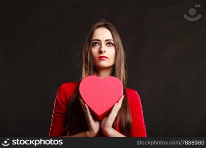 Charity and sharing concept. Young pretty woman in red clothes holding heart box present gift on grey background in studio.