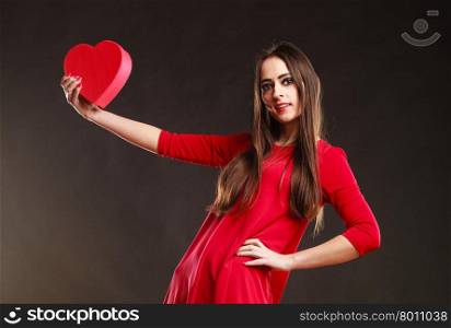 Charity and sharing concept. Young pretty woman in red clothes holding heart box present gift on grey background in studio.